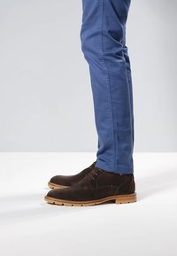 Brune ruskindsankelstøvler med en tanfarvet gummisål, kombineret med blå tætsiddende bukser. Støvlerne har minimal syning og et enkelt design.