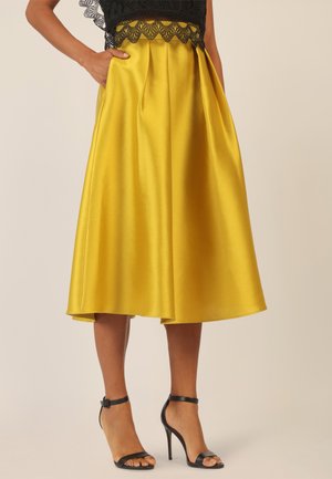Gelber Satinrock mit vollem Design, Falten und seitlichen Taschen, kombiniert mit schwarzen, rückenfreien High-Heels.