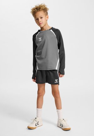 Junges Kind steht in grauer und schwarzer Sportkleidung mit langen Ärmeln und Shorts, trägt weiße Socken und weiße Sportschuhe.