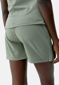 Persona vistiendo pantalones cortos deportivos de color verde oliva y una camiseta a juego, visible de la cintura hacia abajo y por detrás, de pie con la mano relajada al costado.