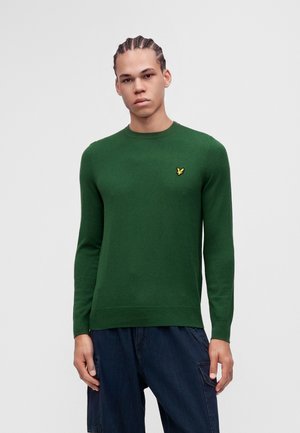 Ung mand med flettet hår bærer en grøn langærmet sweater med en gul fuglelogo og mørkeblå bukser mod en ensfarvet baggrund.