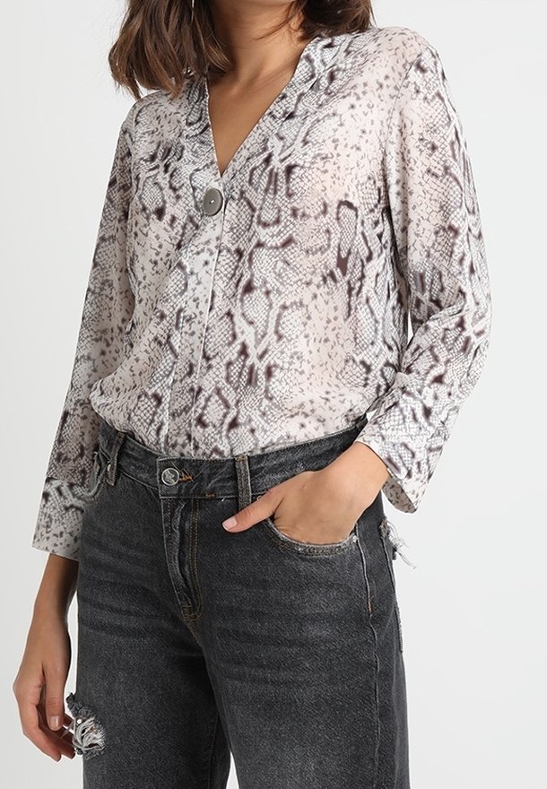 Blouse transparente à imprimé serpent avec un col en V, manches longues et détails à boutons, associée à un jean foncé déchiré. Tons principalement beige et marron.