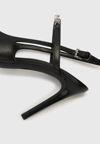 Sandale à talons hauts noire avec une tige en cuir élégante présentant des découpes et des fines lanières autour de la cheville, agrémentée d'une boucle argentée.