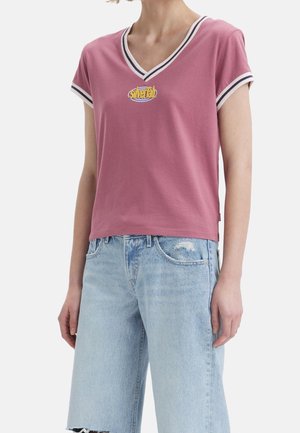 Personne portant un t-shirt rose à col en V avec des bordures blanches et noires et un logo jaune "silverTab", assorti à un short en jean bleu clair déchiré.