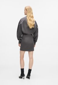 Kvinne med langt blondt h�år iført en grå langermet kjole, svarte sokker og svarte høyhælte sko som står med ryggen til mot en hvit bakgrunn.