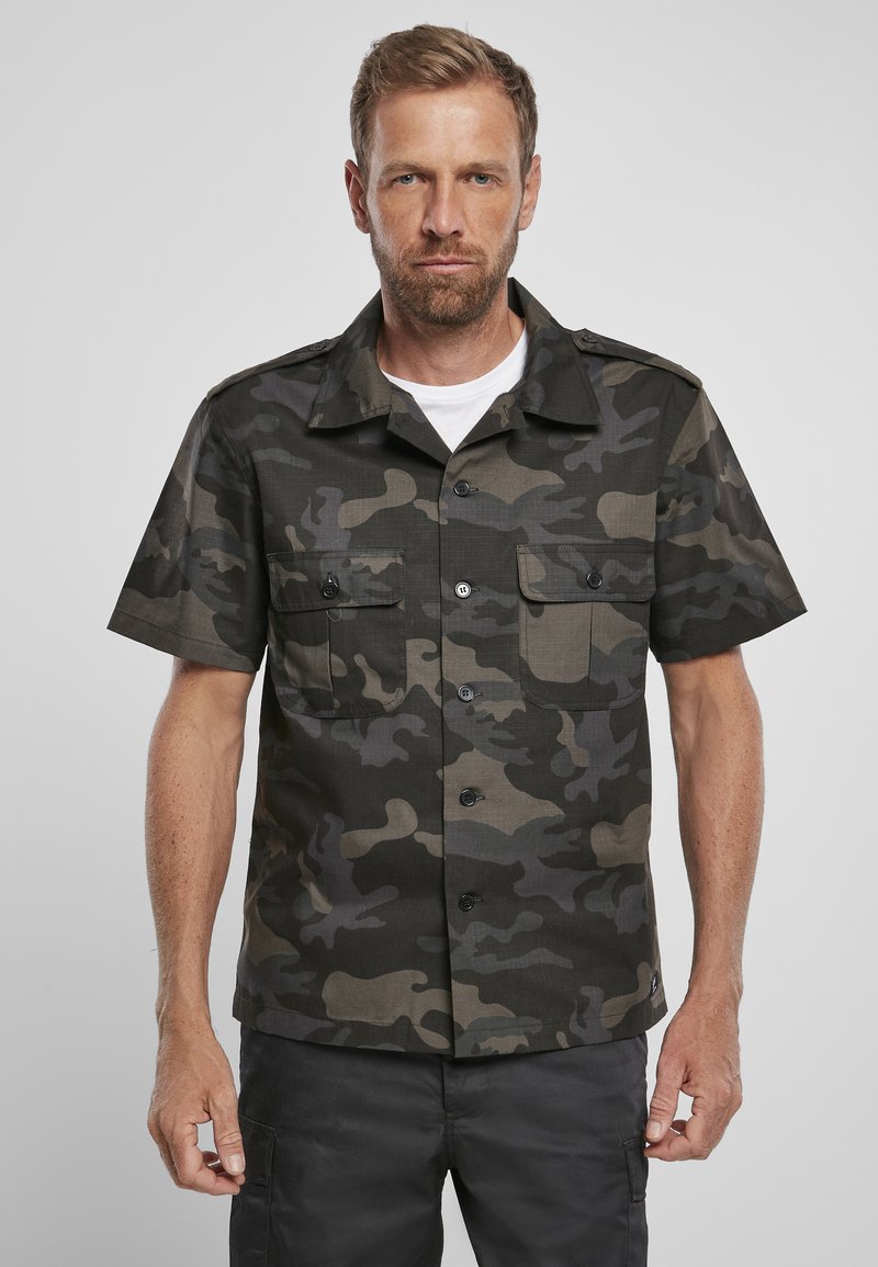 Camicia a maniche corte in camouflage verde scuro e nero. Presenta due tasche sul petto, chiusura con bottoni e colletto a punte. Tessuto texturizzato.