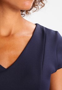 Mörkblå klänning med korta ärmar och V-ringning. Mjukt, stretchigt tyg med subtil lyster. Detaljerad sömnad längs sömmarna.