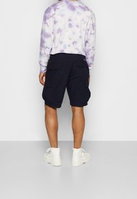 Shorts cargo bleu marine avec grandes poches latérales, associés à un t-shirt à manches longues tie-dye en violet et couleurs pastel, et des baskets montantes blanches.
