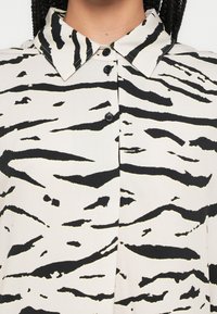 Chemise boutonnée avec col, présentant un motif rayé tigre noir et blanc sur un tissu léger. Quatre boutons noirs visibles.