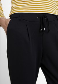 Schwarze Hose mit Kordelzugbund, strukturiertem Stoff, vorderen Falten und Seitentaschen, kombiniert mit einem gelb gestreiften Hemd.