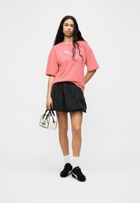 Γυναίκα που στέκεται, φορώντας ροζ μπλουζάκι Puma, μαύρο σορτς, μαύρα αθλητικά Puma, λευκές κάλτσες και κρατώντας μια μικρή λευκή τσάντα Puma.