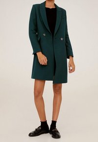 Cappotto in misto lana di colore teal con doppi bottoni, revers a punta e taglio dritto. Presenta tasche e maniche lunghe. Abbinato a scarpe nere.