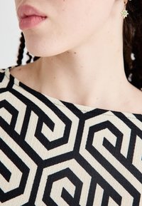 Tielko so vzorom z textúrnej tkaniny s čiernym geometrickým dizajnom na slabo bielej farbe, okrúhly výstrih a viditeľná pokožka pri výstrihu.