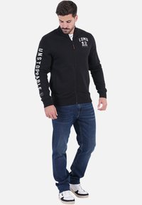 Haut de hoodie zippé noir avec le logo "LOMU" sur la poitrine et le texte "UNSTOPPABLE" sur les manches ; comporte des poignets et un ourlet côtelés, porté avec un jean bleu et des baskets.