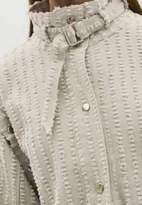 Lähikuva beigeistä teksturoidusta takista, jossa on revittyjä pystysuoria raidoituksia, hopeiset napit ja soljella säädettävä kaulakujanne.