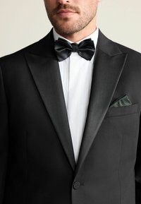 Sacou negru din tuxedo cu revere din satin, cămașă albă cu pliseuri, papion negru și o batistă de buzunar cu un model verde.