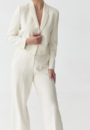 Vrouw gekleed in een op maat gemaakte, crèmekleurige pantalon met een blazer en hooggetailleerde broek, staand tegen een effen lichte achtergrond.