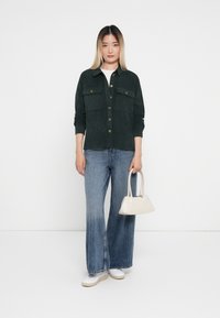 Mørkegrønn kordfløyelskjorte med to brystlommer, knapper foran, kombinert med lyseblå vide jeans og en liten hvit håndveske.