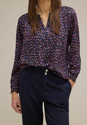 Vrouw draagt een blouse met lange mouwen in paars, zwart en blauw patroon, ingestopt in een high-waisted marineblauwe broek met twee knopen.