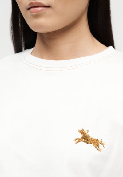 Κοντινό πλάνο ενός ατόμου που φοράει λευκό πουκάμισο με ένα μικρό χρυσό παγιέτα σε σχήμα αλόγου κεντημένο στο στήθος.