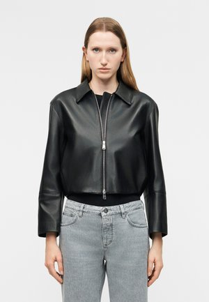 Γυναίκα με ίσια ανοιχτό καστανά μαλλιά, φοράει μαύρο δερμάτινο κοντομάνικο μπουφάν με φερμουάρ πάνω από μαύρο τοπ και ανοιχτό γκρι τζιν, στέκεται μπροστά σε λευκό φόντο.