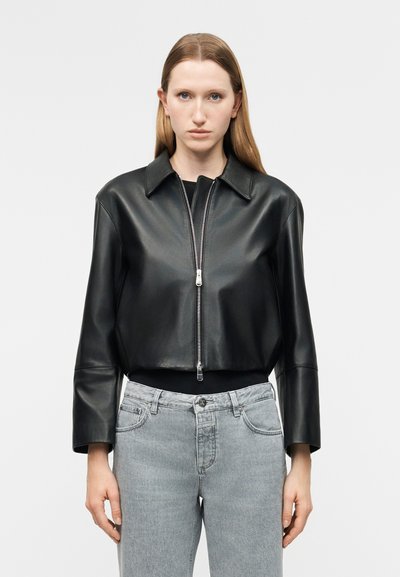 Femme aux cheveux bruns clairs et raides portant une veste en cuir noire courte fermée par une fermeture éclair sur un haut noir et un jean gris clair, debout devant un fond blanc.