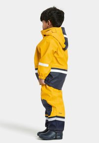 Heldere gele waterdichte jas en broek met marineblauwe accenten, voorzien van reflecterende strepen en elastische manchetten, gecombineerd met zwarte rubberen laarzen.