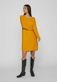 Vestito a maglia giallo senape a maniche lunghe con una vestibilità morbida. Collo e orlo a costine. Abbinato a stivali neri in pelle al ginocchio.