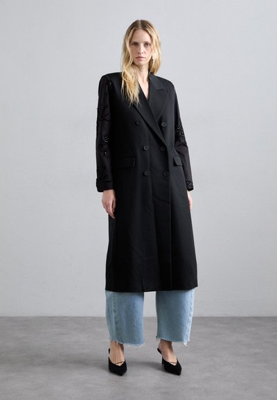 Abrigo negro de doble botonadura con mangas largas, que presenta una tela transparente y adornos, combinado con unos vaqueros de pierna ancha en azul claro y tacones negros.