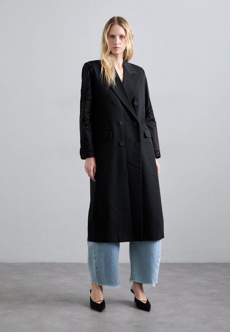 Melnā dubultā jaku ar garām piedurknēm, kas izgatavota no caurspīdīga auduma un rotāta ar dekoratīviem elementiem, apvienota ar gaiši zilām platajām biksēm un melniem papēžiem.