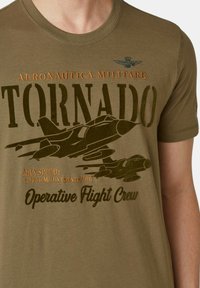 T-shirt di cotone verde oliva con una grafica di jet da combattimento, impreziosita dal testo "TORNADO" e dai dettagli dell'equipaggio di volo in nero e arancione a contrasto.