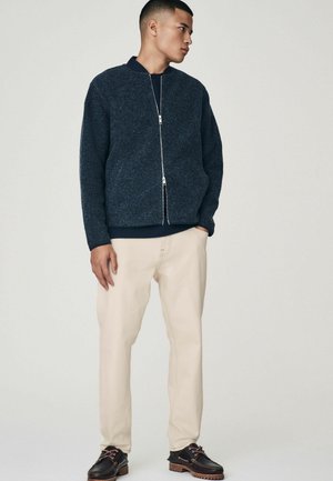 Chaqueta bomber de cremallera azul oscuro en un tejido texturizado, combinada con pantalones beige claros y zapatos oscuros con acentos marrones.
