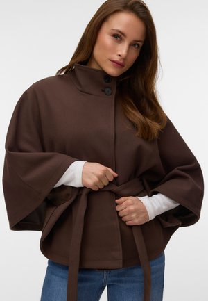 Femme aux longs cheveux bruns attachant la ceinture d’un manteau cape brun foncé par-dessus un haut blanc à manches longues et un jean bleu.