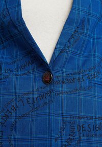 Tissu de chemise à carreaux bleus avec un motif texturé, comprenant un bouton noir près du col et un texte imprimé en encre foncée.