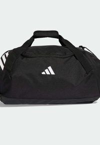 Bolsa de viaje Adidas negra con asa acolchada, logo blanco de Adidas en la parte frontal y diseño parcial de tres rayas en el lateral sobre un fondo blanco.