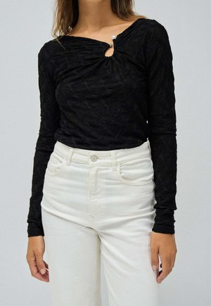 Femme portant un haut noir texturé à manches longues avec un détail en perles au niveau de l'encolure et un jean blanc taille haute sur un fond uni.