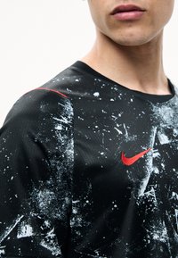 Νεαρό άτομο φοράει μια σπορ μπλούζα μαύρου και λευκού χρώματος με πιτσιλιές, με κόκκινο λογότυπο Nike στο στήθος, περικομμένη στον λαιμό και τον ώμο.