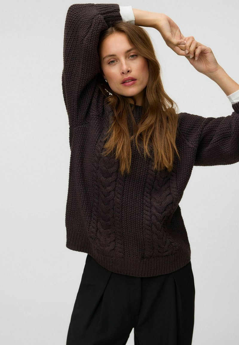 Femme aux longs cheveux bruns portant un pull en maille torsadée foncé et un pantalon noir, posant avec les bras levés sur un fond uni.