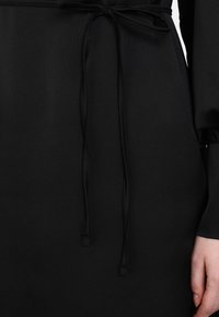 Black dress featuring a smooth, textured fabric. Detail includes a tied belt at the waist and long sleeves, with a straight cut silhouette.