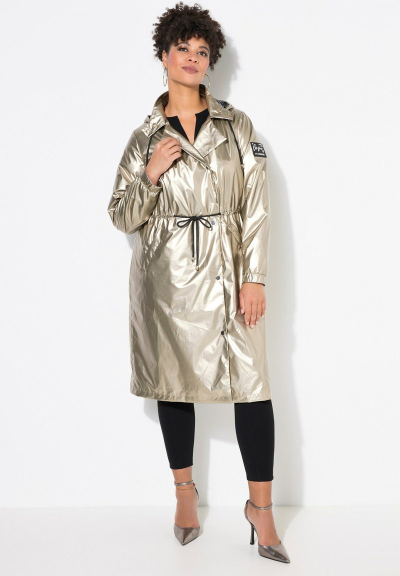 Metallisch gouden regenjas met een ceintuur in de taille, knoopsluiting en zijzakken. Voorzien van een standaard kraag en elastische manchetten.