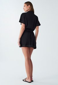 Donna che indossa un vestito corto nero con maniche corte, in piedi a piedi nudi su uno sfondo chiaro e semplice, rivolta di lato.