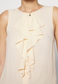 Helles pfirsichfarbendes ärmelloses Blusenshirt mit einem geschichteten Rüschendesign aus weichem Material und einem kleinen dekorativen Reißverschlussverschluss am Ausschnitt.