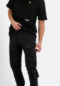 Zwarte cargobroek met meerdere met ritsen gesloten zakken, gemaakt van stevig materiaal. Draag het met een bijpassend zwart T-shirt met een logo op de voorkant.