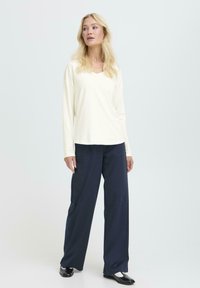 Bluză cu mânecă lungă și decolteu în V, dintr-un material alb crem, asortată cu pantaloni largi bleumarin. Modelul poartă pantofi negri lucioși, cu un design minimalist, fără modele.