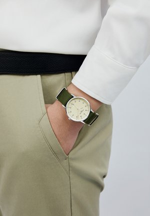 Silver watch with a round face, cream dial, black markings, and colorful accents. Green fabric strap attached to a wrist in a pocket.