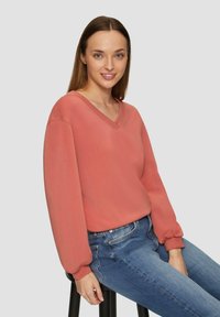 Koraalrode V-hals pullover van zachte stof met lange, gathered mouwen, gecombineerd met aansluitende blauwe jeans. Eenvoudig en ontspannen ontwerp.