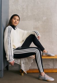 Pulôver branco felpudo com riscas pretas, leggings pretas com riscas brancas e sapatos prateados. Modelo posa em um banco de madeira contra uma parede de concreto.