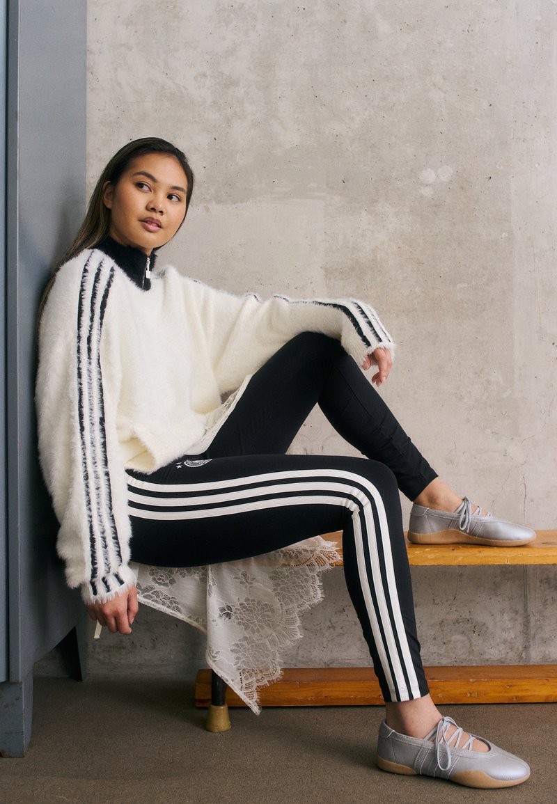 Pulôver branco felpudo com riscas pretas, leggings pretas com riscas brancas e sapatos prateados. Modelo posa em um banco de madeira contra uma parede de concreto.