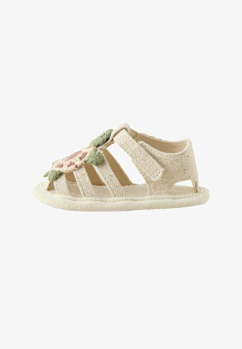 Sandale beige pour tout-petit avec des lanières en tissu doux, une fermeture à scratch et une application de fleur rose et verte sur le dessus.