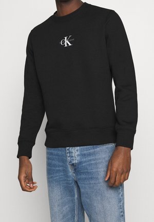Homme portant un sweat-shirt noir Calvin Klein avec un logo blanc et un jean bleu clair, se tenant devant un fond uni.